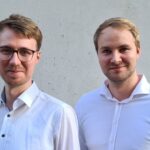 von Tobias Greiner links und Oliver Greiner rechts, in hellblauen Hemden stehen lächelnd vor einer hellgrauen Wand.